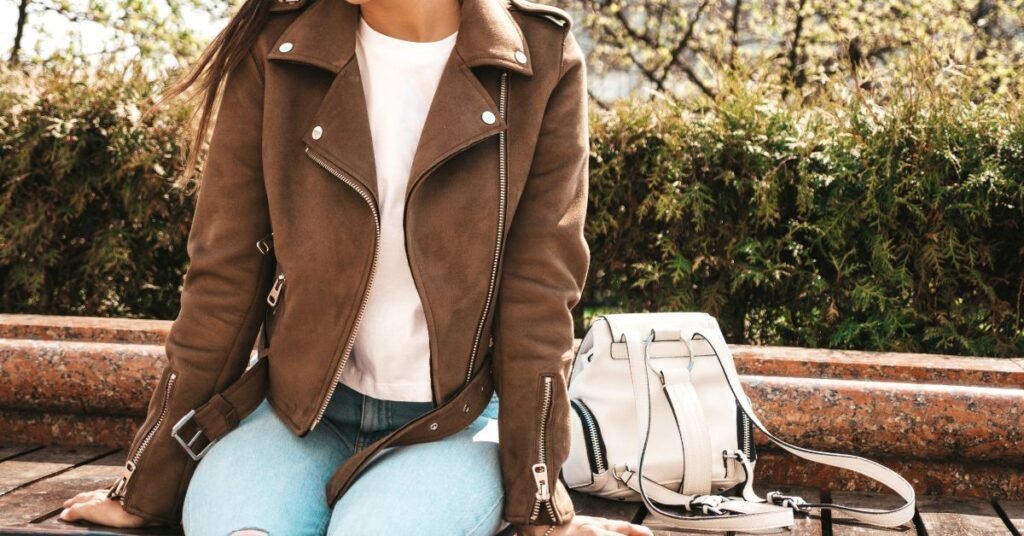 Femme souriante en perfecto marron, lunettes roses et sac blanc, exemple d’un look cuir féminin et stylé